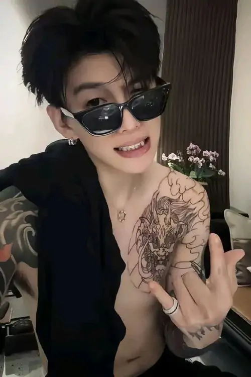 32 Chàng trai đeo kính đen, mặc áo mở sâu và khoe hình xăm lớn trước gương, tay giơ ký hiệu đầy tự tin, bức ảnh mang năng lượng nổi bật, có độ nóng bỏng rõ nhưng vẫn giữ được sắc thái phong độ và tự chủ